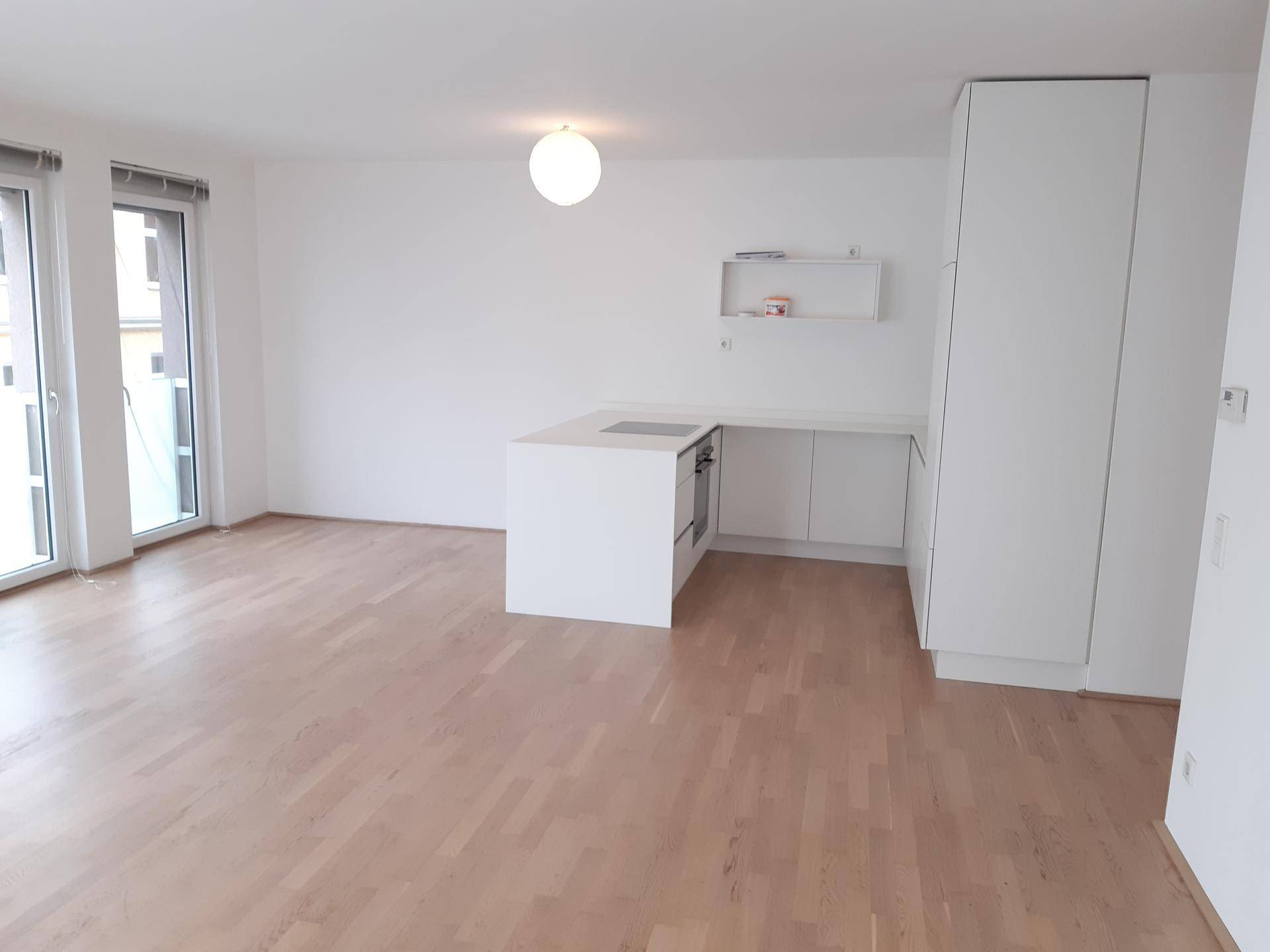 Offener Wohnbereich mit integrierter weißer Küche, Holzboden und viel Tageslicht durch große Fenster.