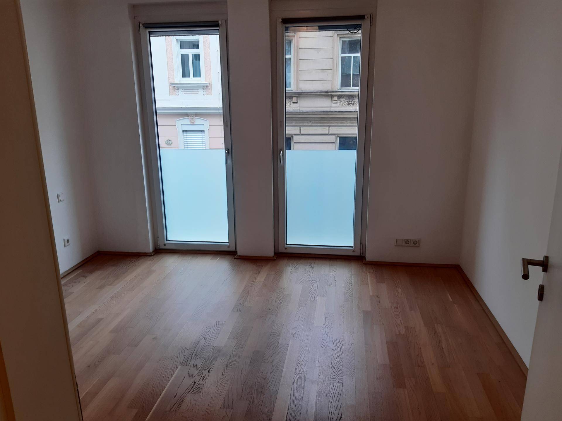 Helles Schlafzimmer mit Holzboden und zwei großen Fenstern, die einen Blick auf die Stadt bieten.
