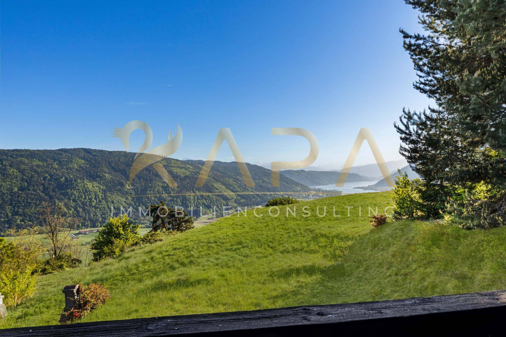 Weitläufiger Blick über grüne Hügel und Täler bis zum See unter einem klaren blauen Himmel.