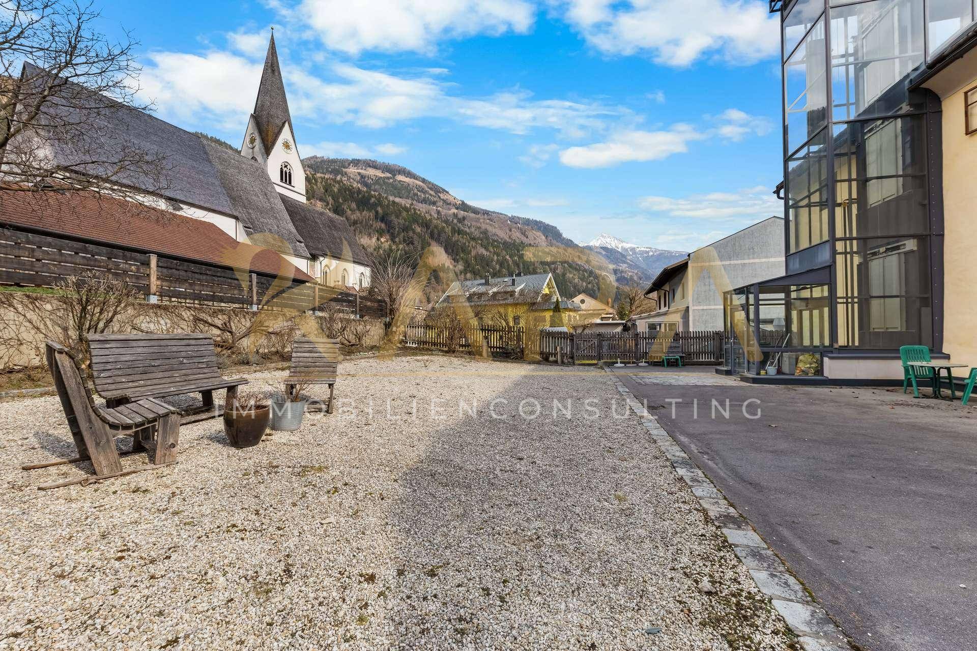 Freie Kiesfläche neben einem Gebäude mit Kirche und Bergen im Hintergrund.