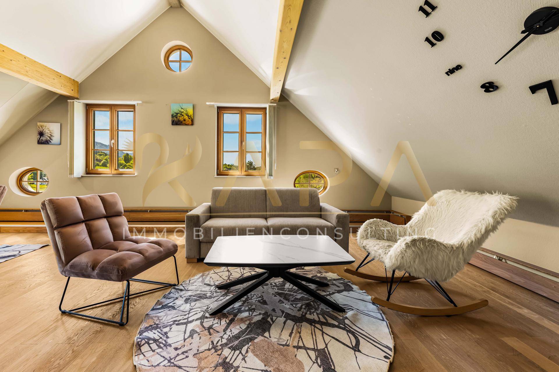 Gemütliches Wohnzimmer im Dachgeschoss mit Holzbalken, runden Fenstern und modernem Mobiliar.