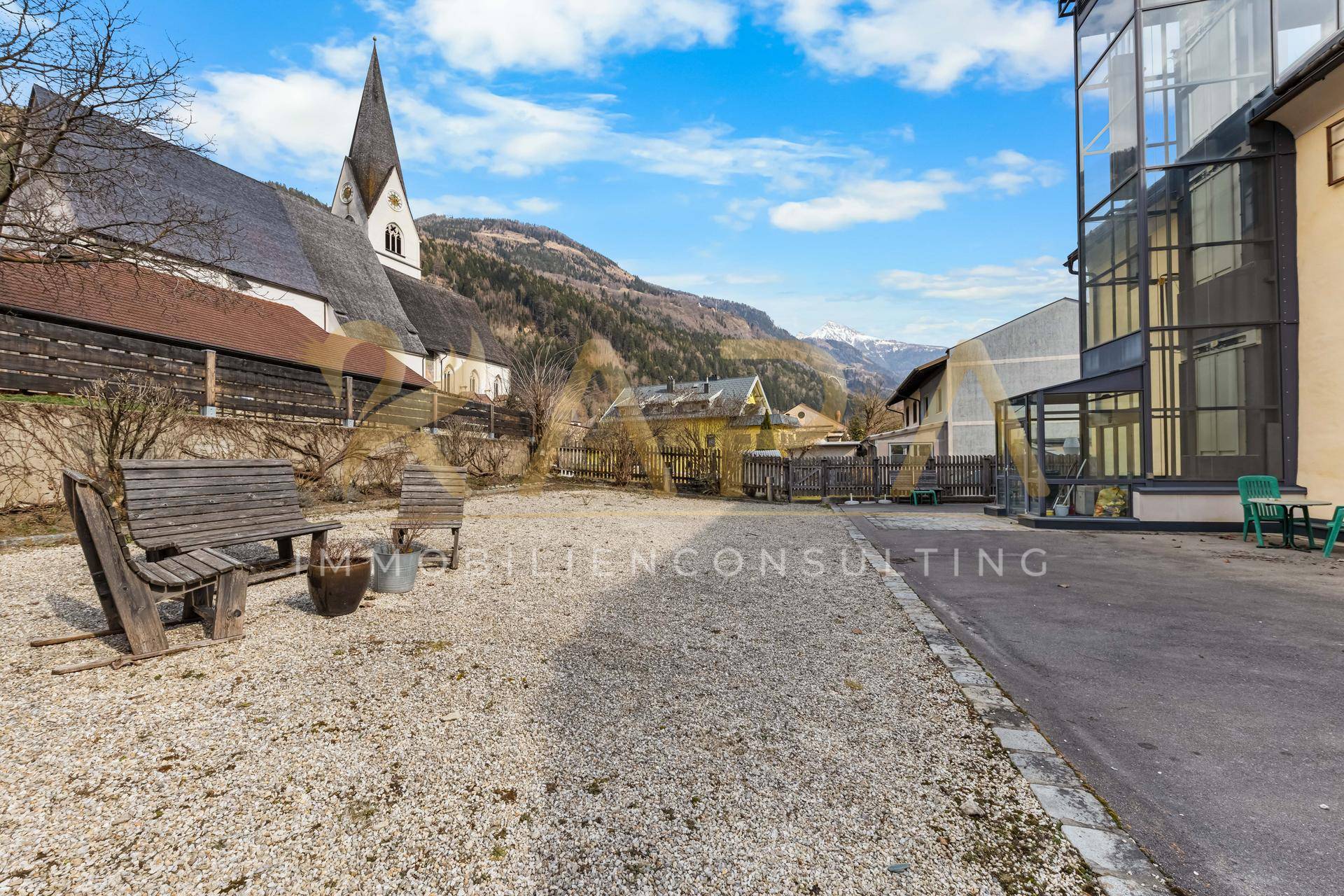 Außenbereich mit Kiesfläche, Holzbänken und Blick auf die Berge und eine Kirche.