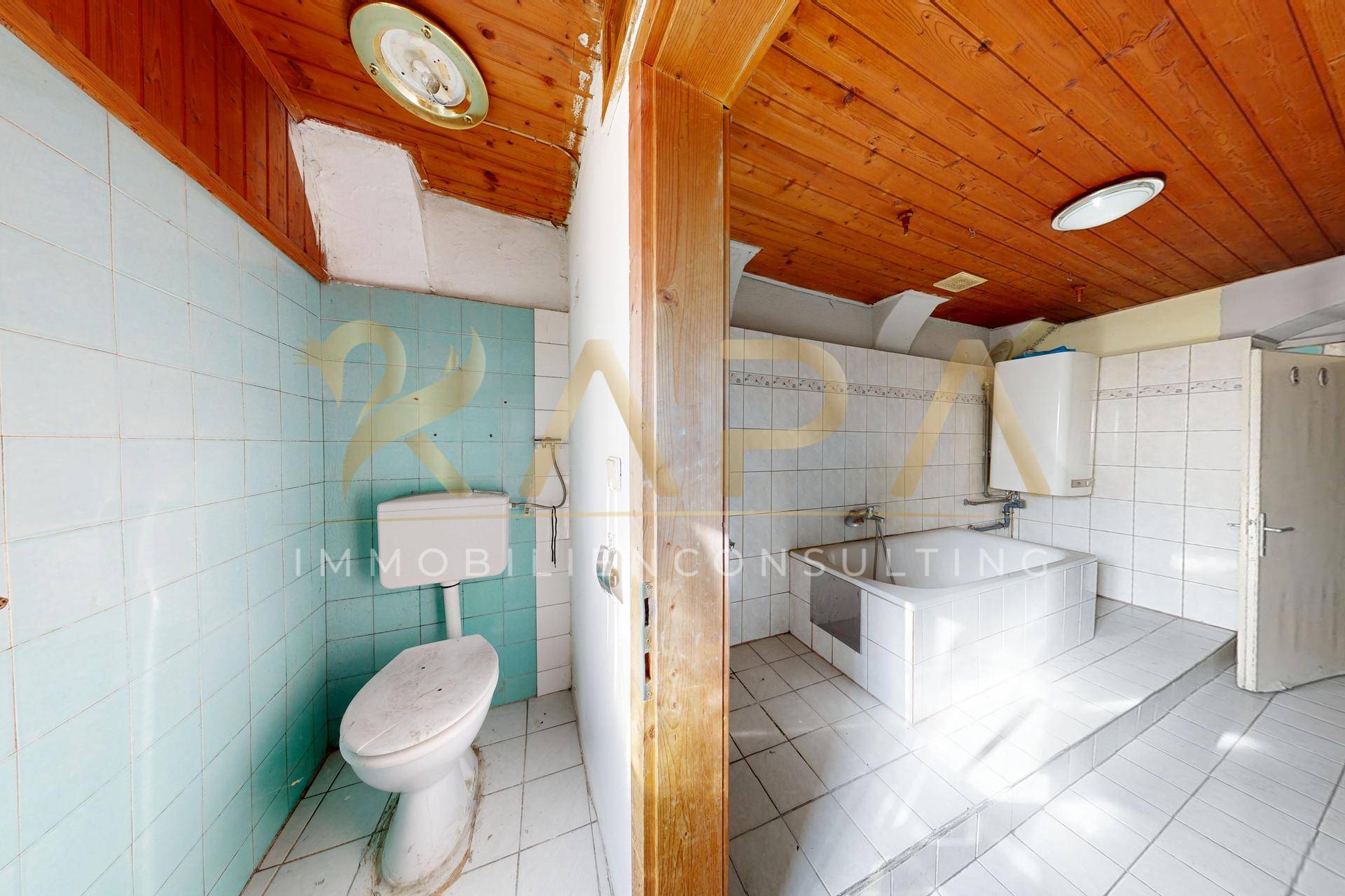 Badezimmer mit blauen Fliesen, Toilette, Badewanne und Holzdecke, renovierungsbedürftig.