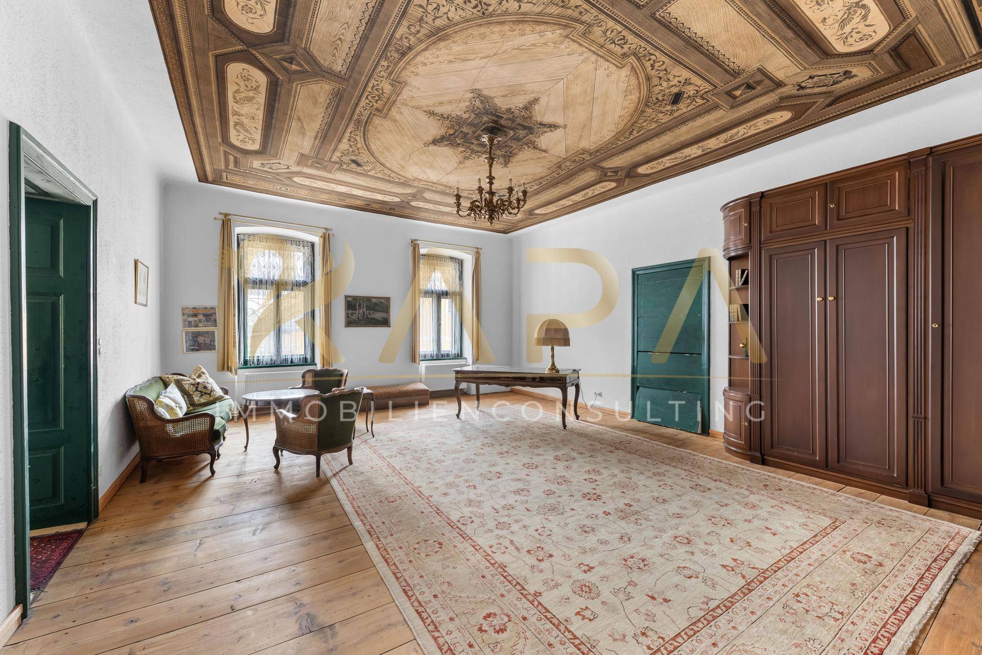 Geräumiges Wohnzimmer mit kunstvoll bemalter Kassettendecke, Holzdielenboden und großem Teppich, klassisch möbliert.