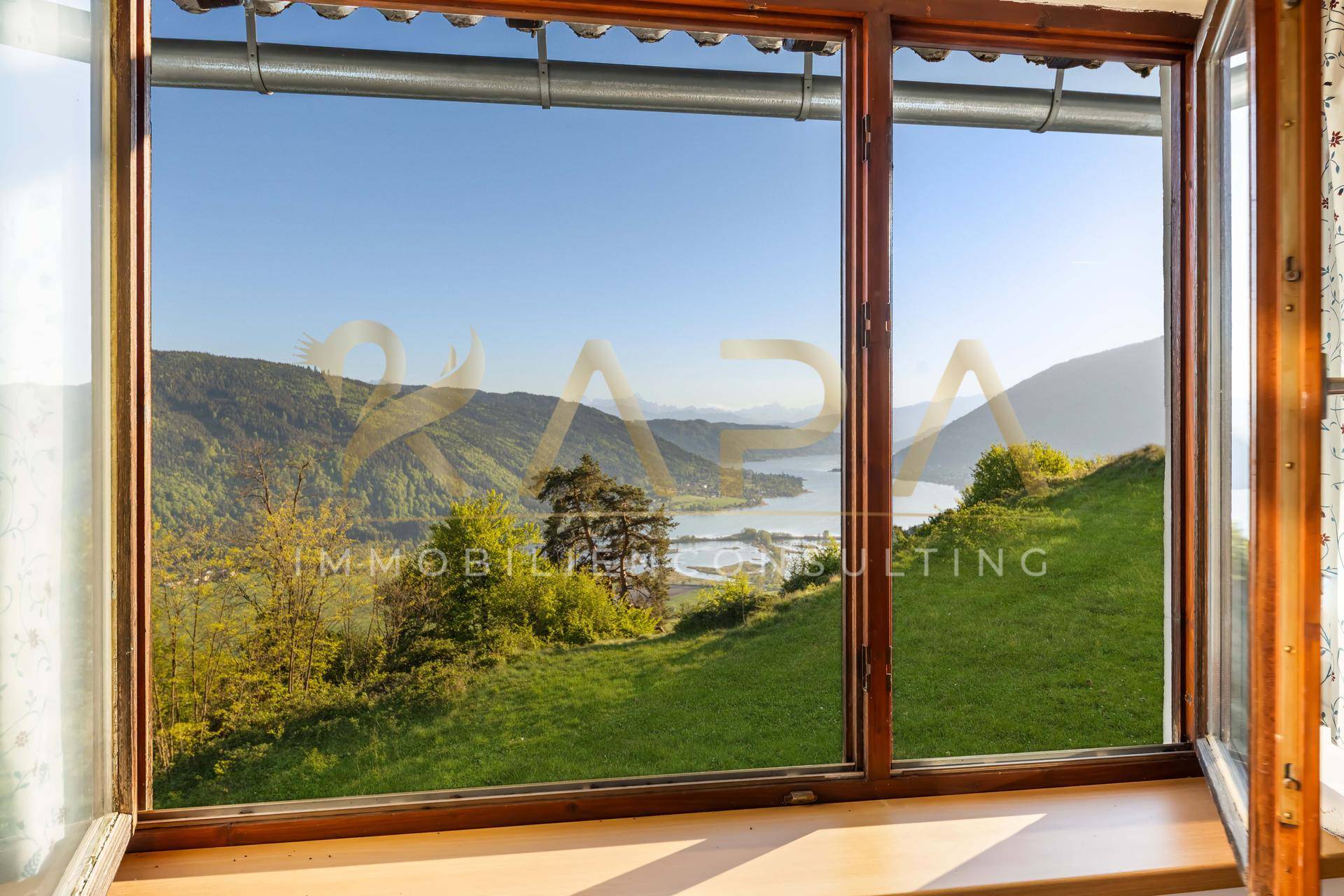 Fensterblick auf die malerische Berglandschaft mit See und grünen Wiesen unter strahlend blauem Himmel.