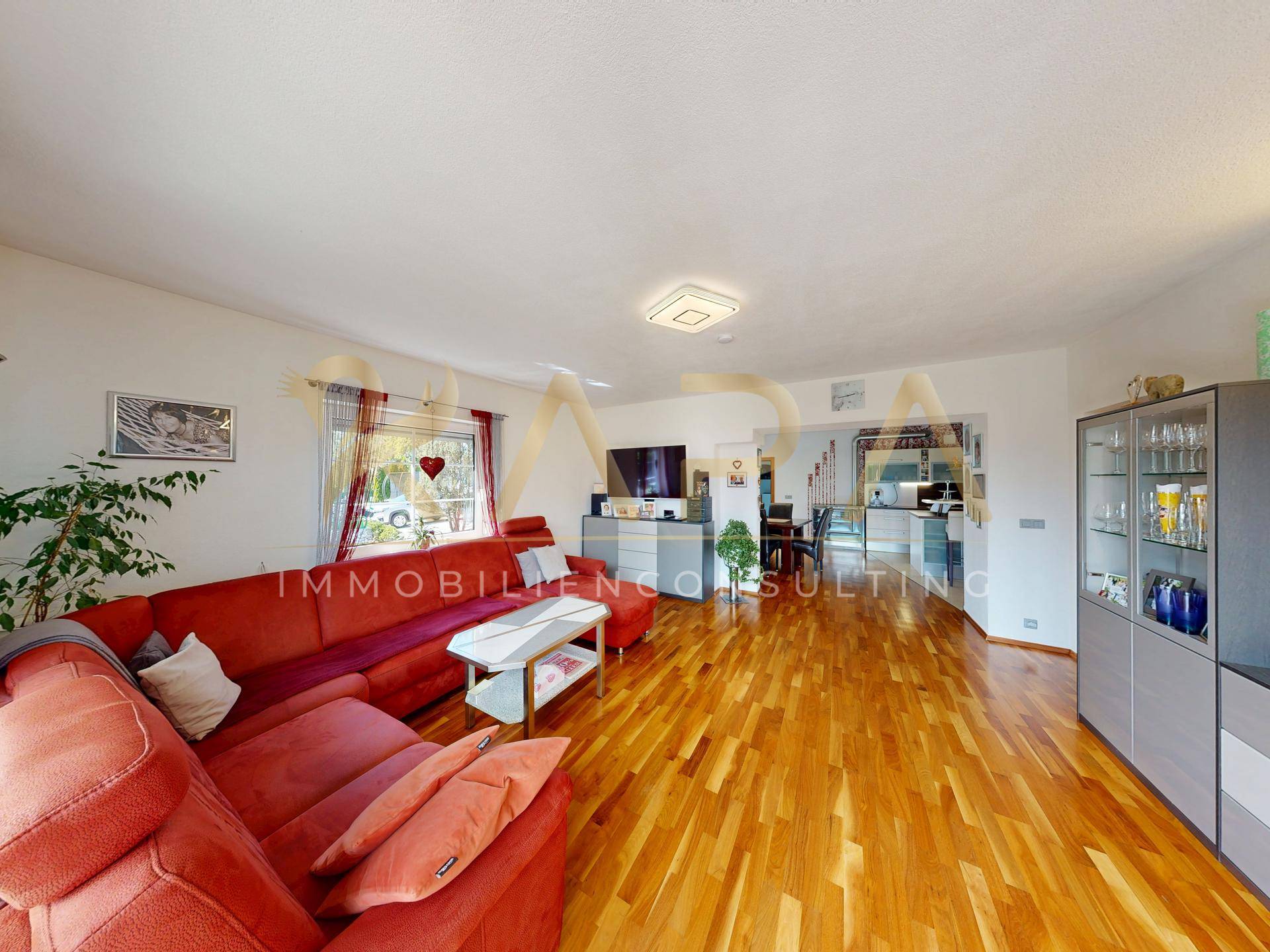 Helles Wohnzimmer mit rotem Sofa, Holzboden und offenem Übergang zum Essbereich und zur Küche.