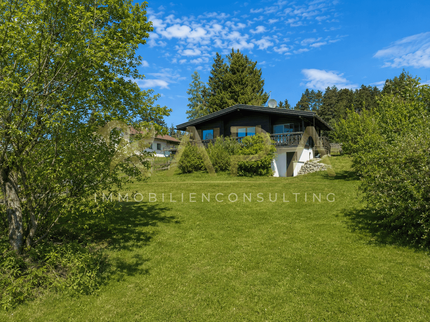 Holzhaus mit Balkon und Gartenbereich, umgeben von viel Grün und Bäumen.