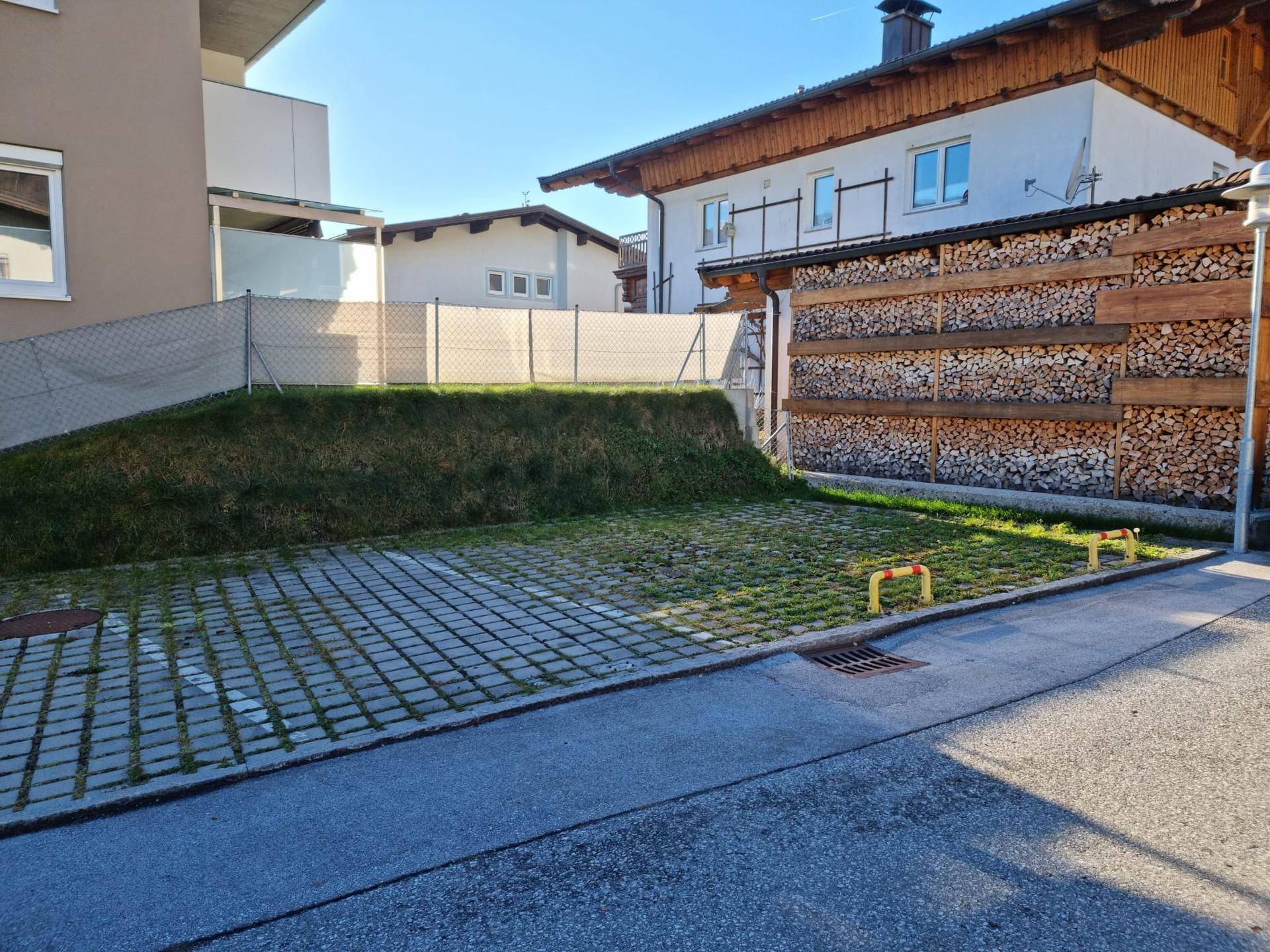 Begrünter Parkplatzbereich vor einem modernen Gebäude mit angrenzendem traditionellen Haus und Holzstapel.