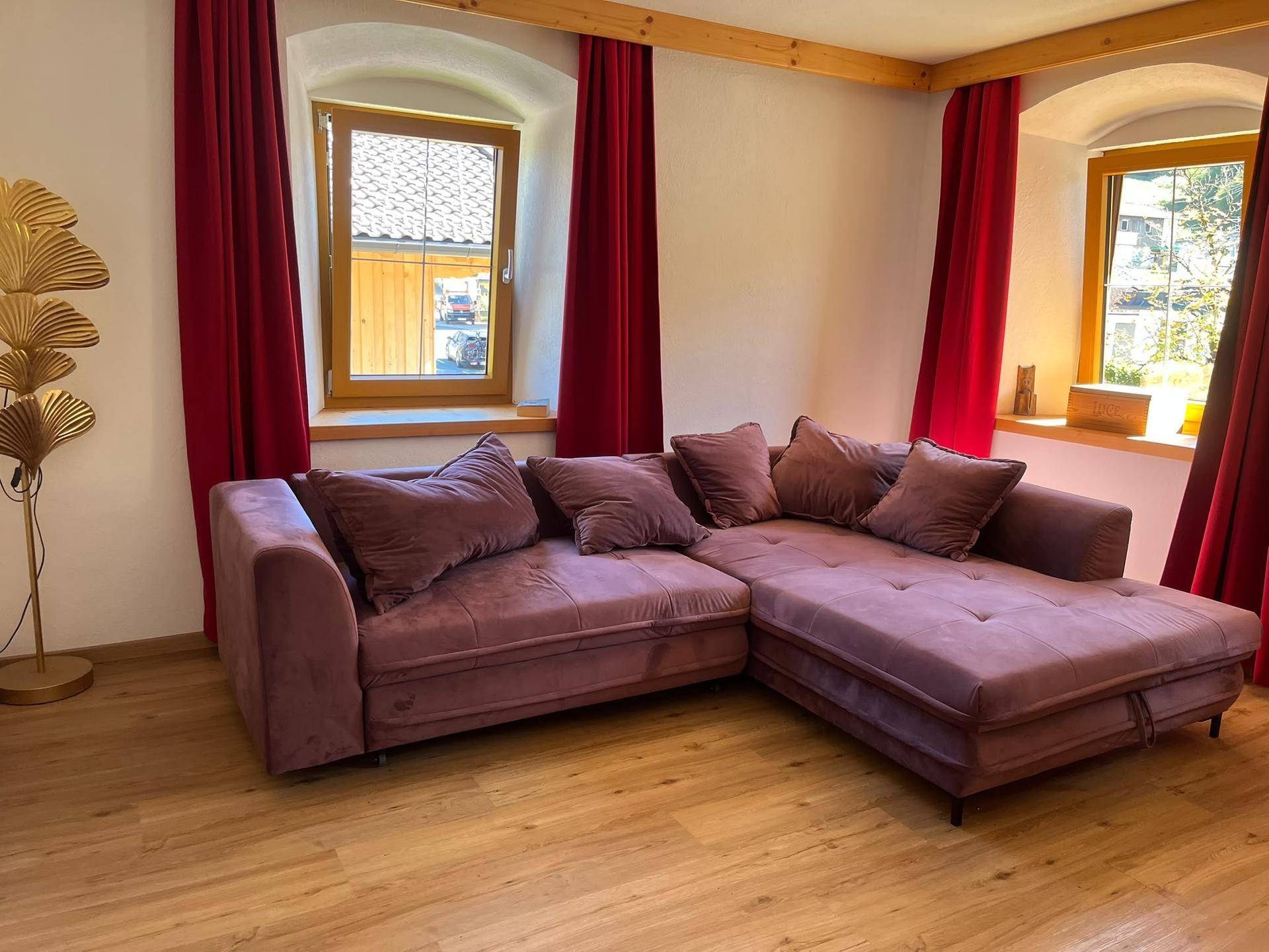 Gemütliches Wohnzimmer mit großem Ecksofa, Holzboden und Fenstern mit roten Vorhängen.