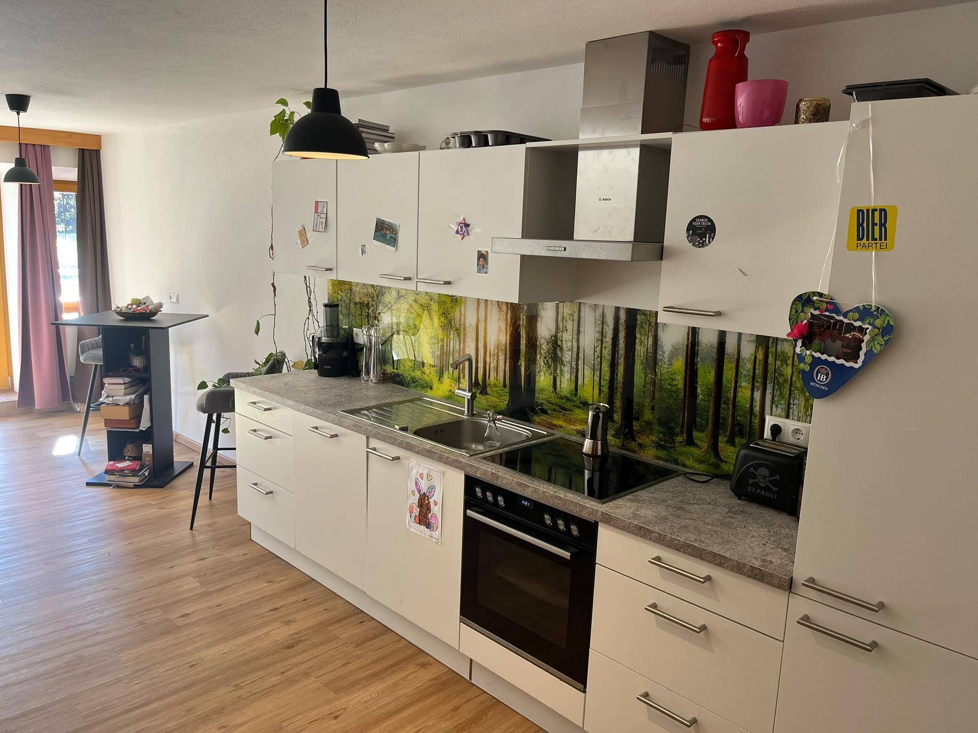 Helle Einbauküche mit weißen Fronten, integrierten Geräten und dekorativer Wald-Rückwand.