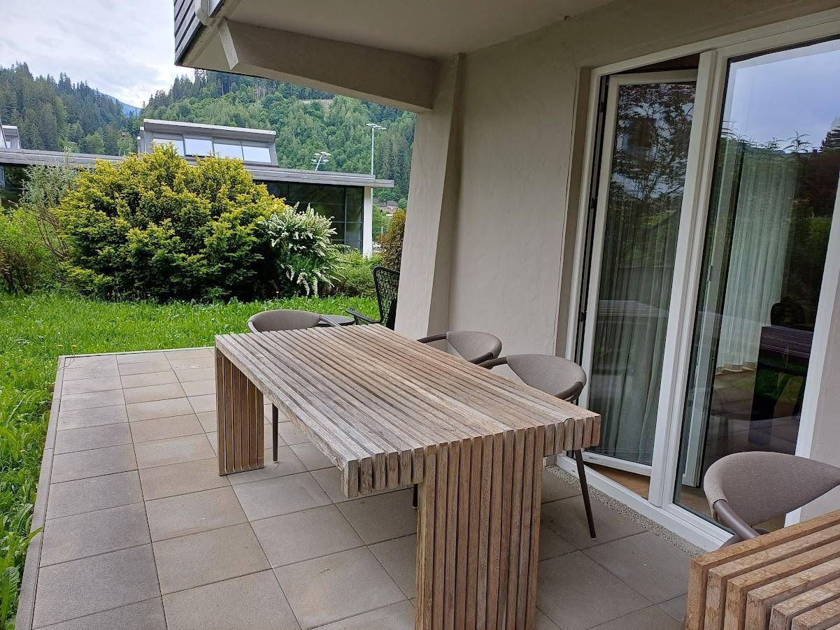 Sonniger Terrassenbereich mit Holztisch und Stühlen, umgeben von Grünflächen und Bergblick.