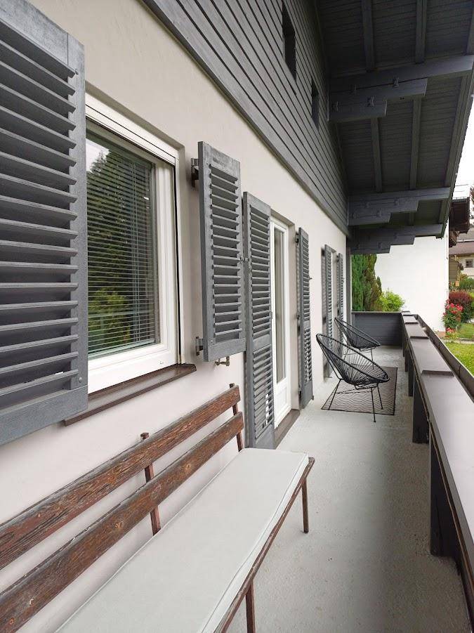 Langer Balkon mit Holzsitzbank, modernen Stühlen und grauen Fensterläden mit Blick ins Grüne.