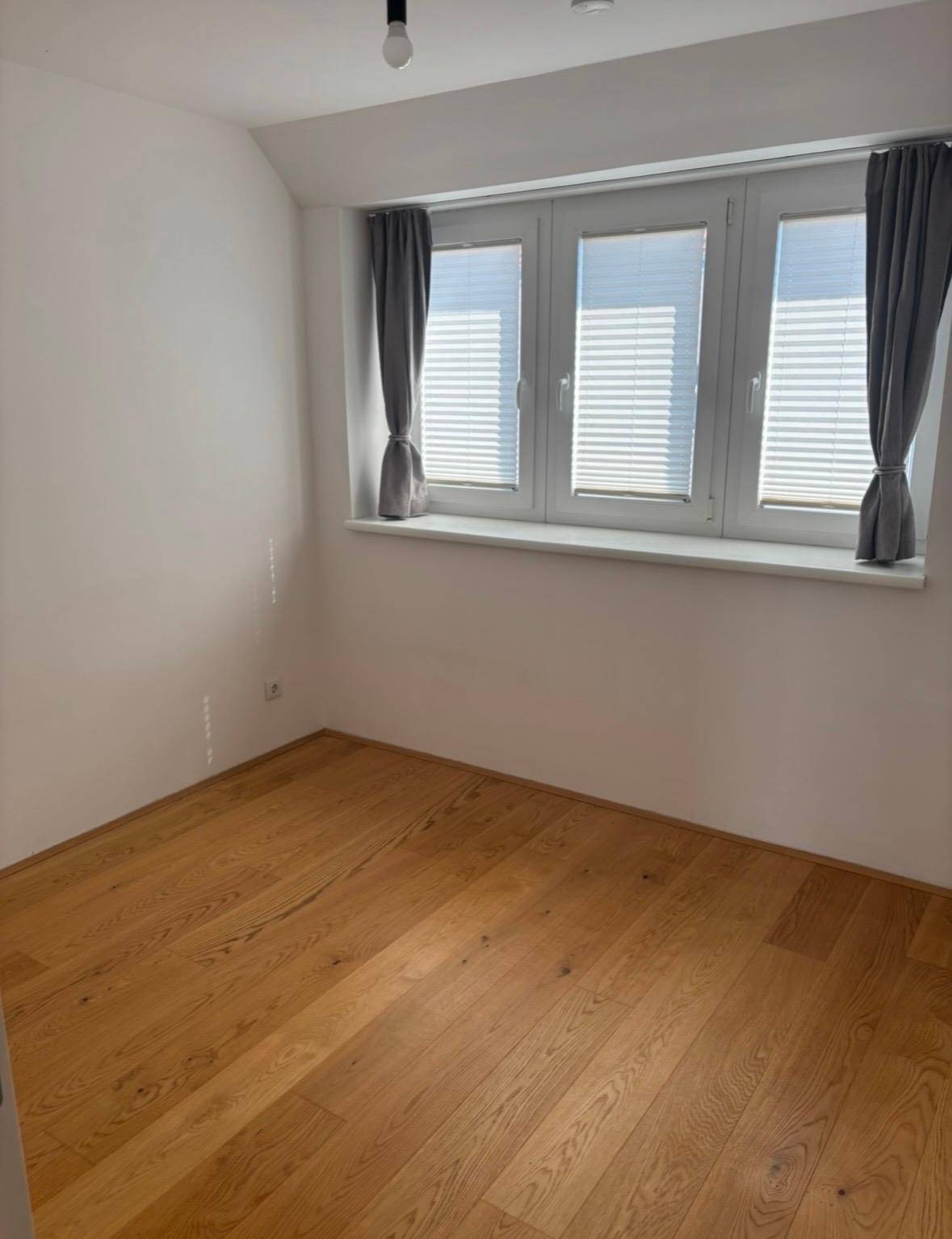 Leerer Raum mit Holzboden und einem Fenster mit grauen Vorhängen, das viel Licht bietet.