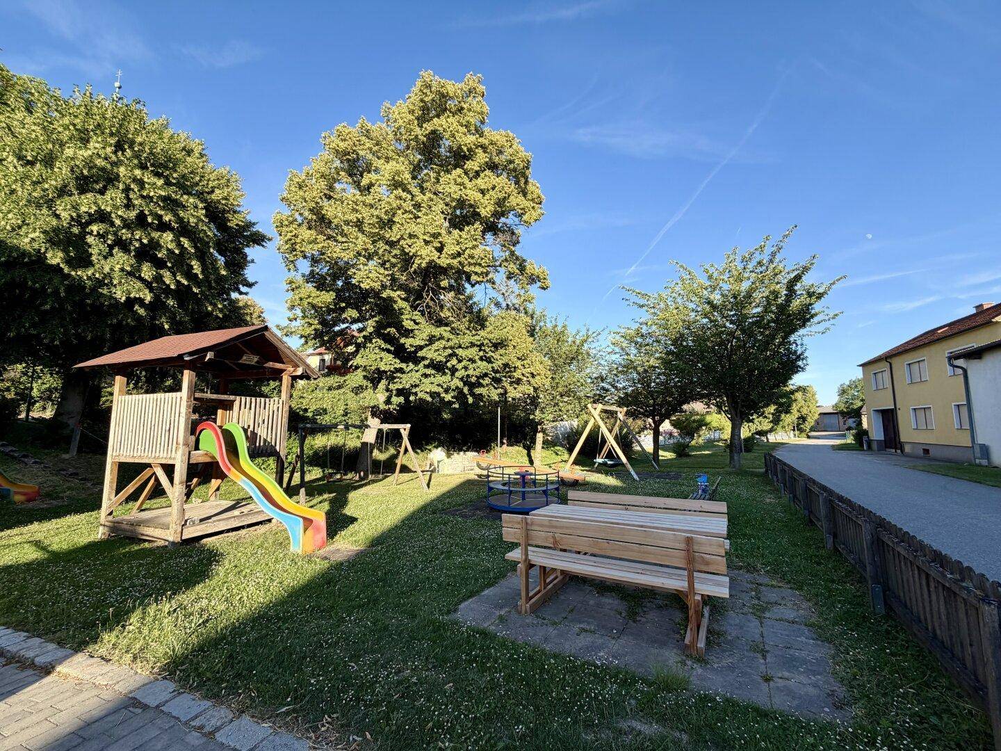 Sonniger Kinderspielplatz mit Rutsche, Schaukeln und Bänken in einer Wohngegend.