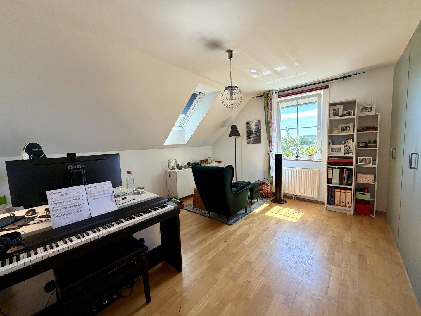 Vielseitiger Raum im Dachgeschoss mit Klavier, Lesesessel, Bücherregal und Dachfenster.