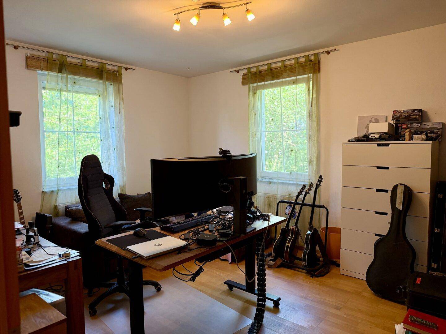 Helles Zimmer mit zwei Fenstern, Schreibtisch und Musikinstrumenten, ideal als Schlaf- oder Arbeitszimmer.