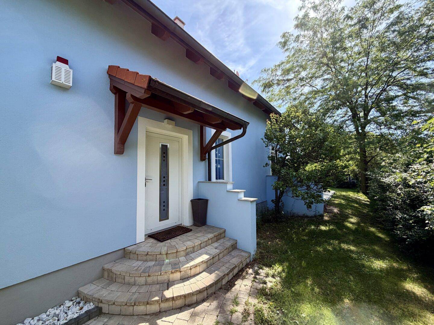 Eingangsbereich des Hauses mit weißer Tür, Steinstufen und einem schützenden Holzvordach.