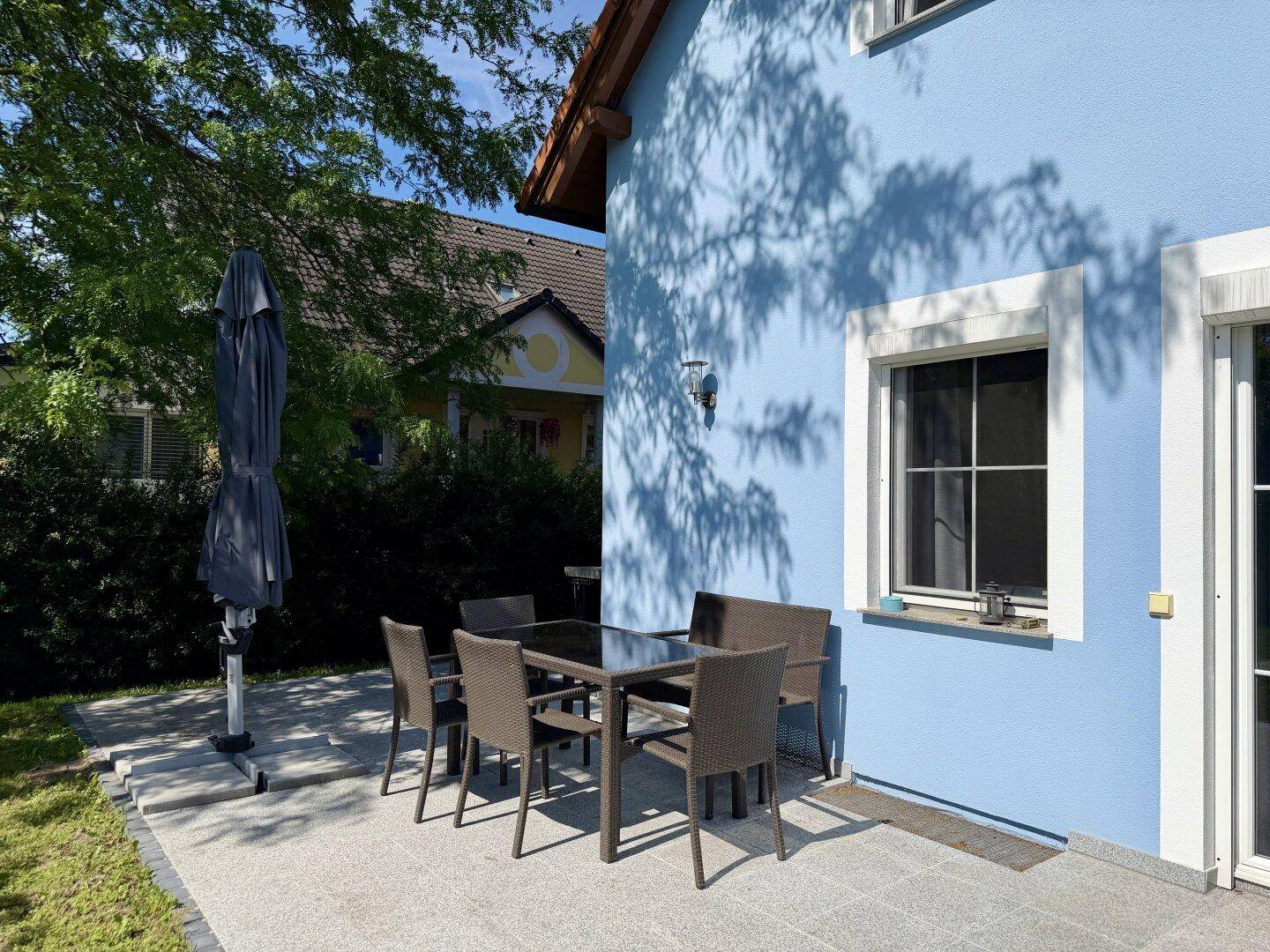 Sonniger Terrassenbereich mit Esstisch und Stühlen neben der blauen Hausfassade.