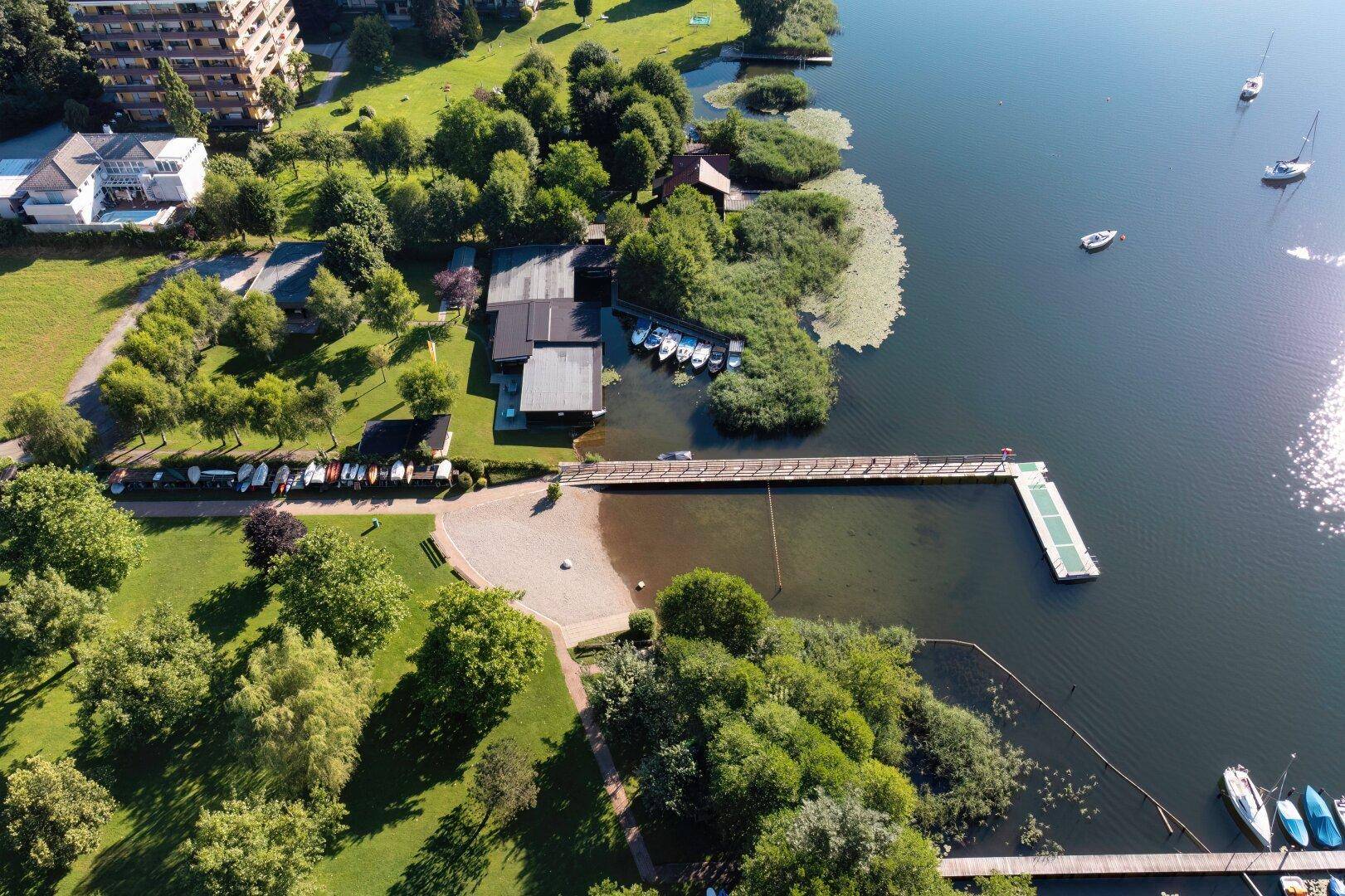 Luftaufnahme eines Sees mit Uferbebauung, grünen Bäumen, Badestrand und langen Bootsstegen.