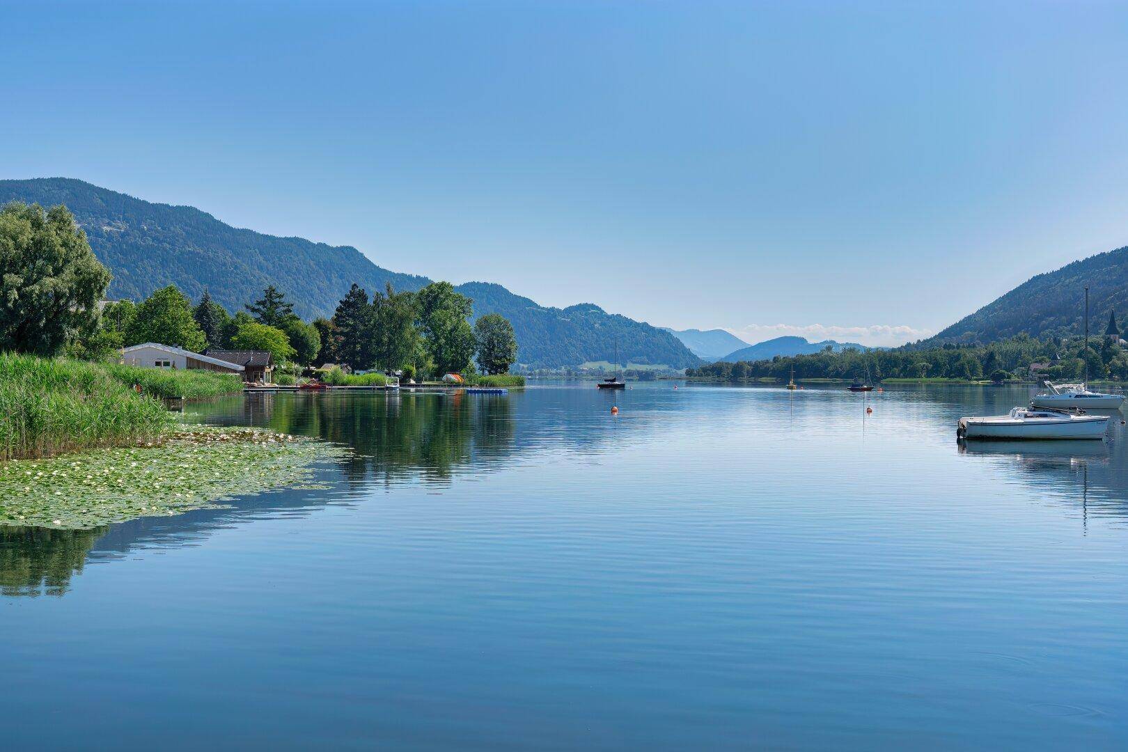 Weitläufiger Seeblick mit ruhigem Wasser, umgeben von bewaldeten Hügeln unter blauem Himmel.