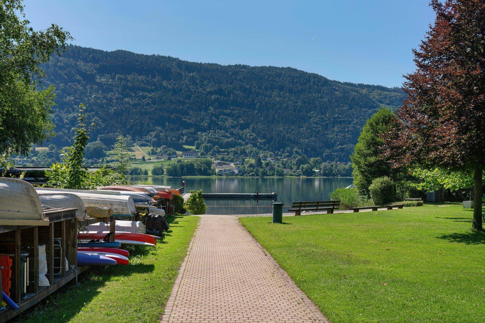 Gepflasterter Weg führt zum Seeufer, gesäumt von Grünflächen, Bäumen und gelagerten Booten.