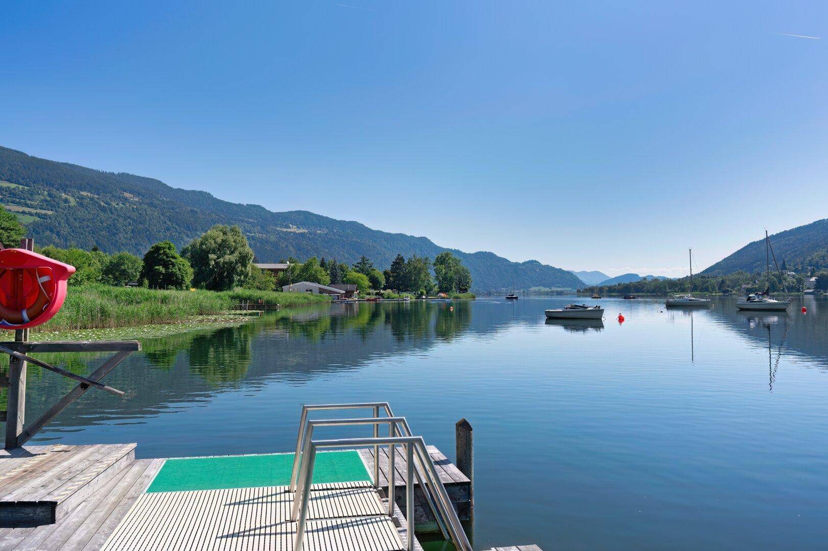 Weitläufiger Seeblick von einem Holzsteg mit Badeleiter, umgeben von Bergen und Booten.