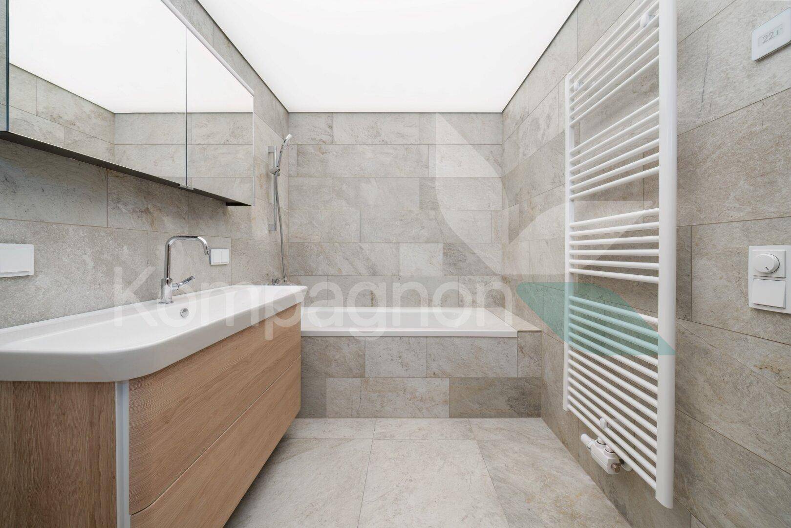 Modernes Badezimmer mit grauen Fliesen, Holzwaschtisch, Badewanne und beheiztem Handtuchhalter.