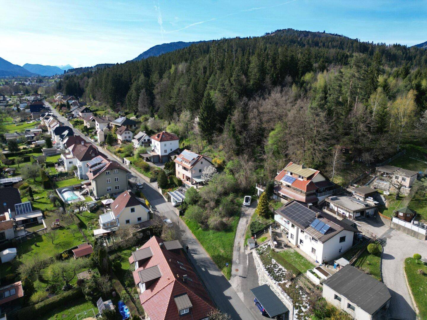 Wohnstraße mit Einfamilienhäusern, einigen Solaranlagen und angrenzendem Waldgebiet.