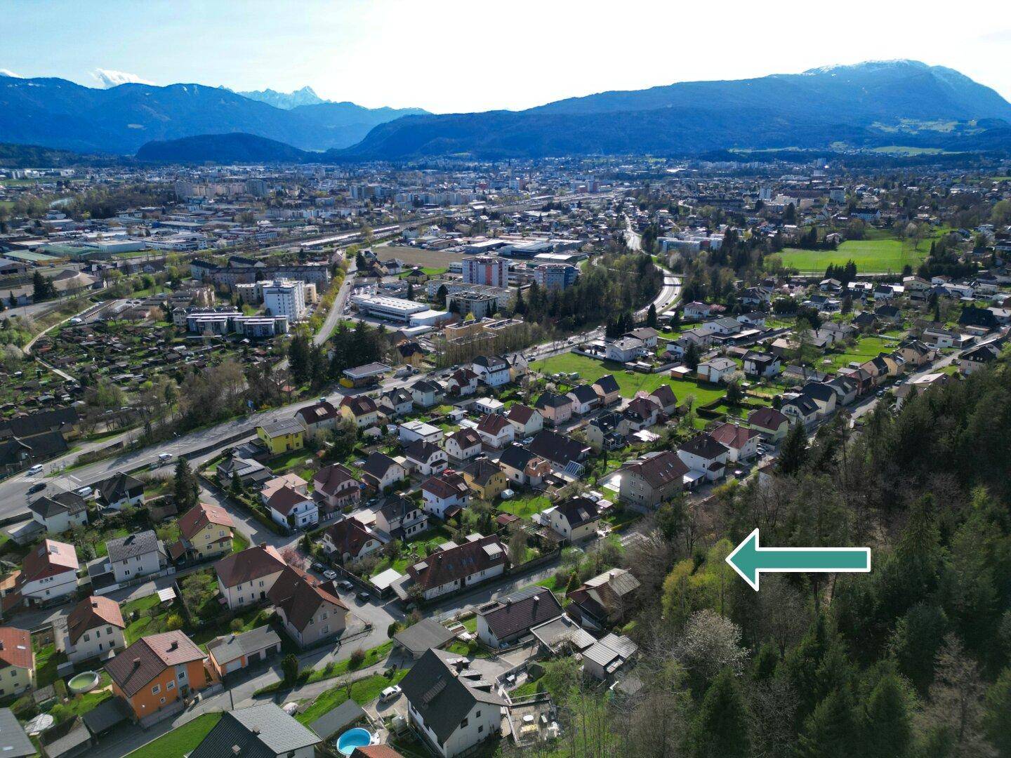 Panoramische Luftaufnahme einer Stadtlandschaft mit Wohngebieten, Wald und Bergketten am Horizont.