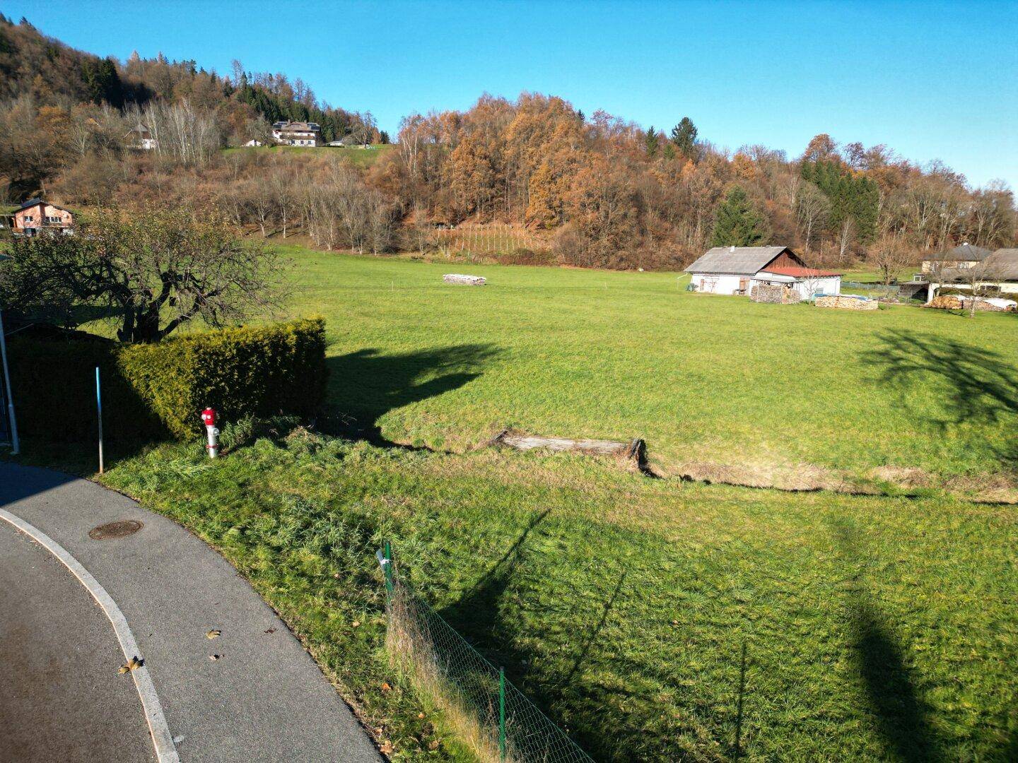 Weitläufige grüne Wiese mit einem kleinen Bachlauf, umgeben von Bäumen und einigen Häusern in der Ferne.