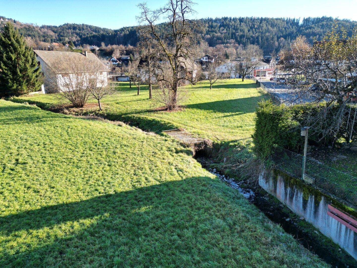 Sonnige grüne Wiese mit einem kleinen Bachlauf und alten Bäumen, umgeben von ländlicher Bebauung.