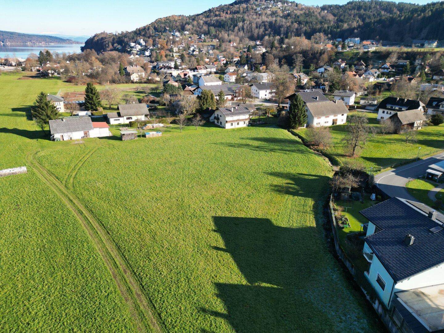 Panoramablick über eine weite grüne Wiese mit Häusern und einem See in der Ferne unter blauem Himmel.