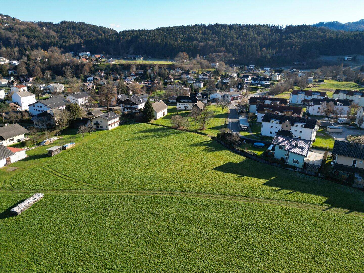 Weitläufige Luftaufnahme einer grünen Wiese mit umliegenden Häusern und bewaldeten Hügeln unter blauem Himmel.