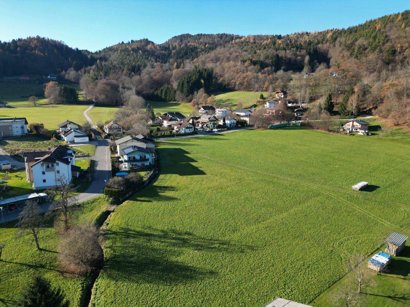 Weitläufige Luftaufnahme einer grünen Wiese mit umliegenden Häusern und bewaldeten Hügeln unter blauem Himmel.