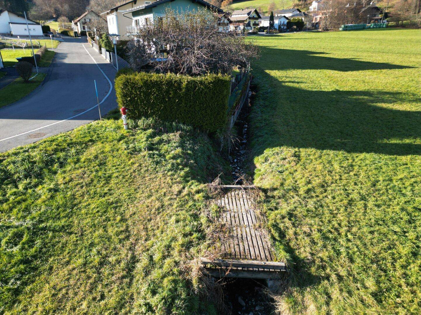 Grüne Wiese mit einem kleinen Bachlauf und einer Holzbrücke, angrenzend an eine Straße und eine Hecke.
