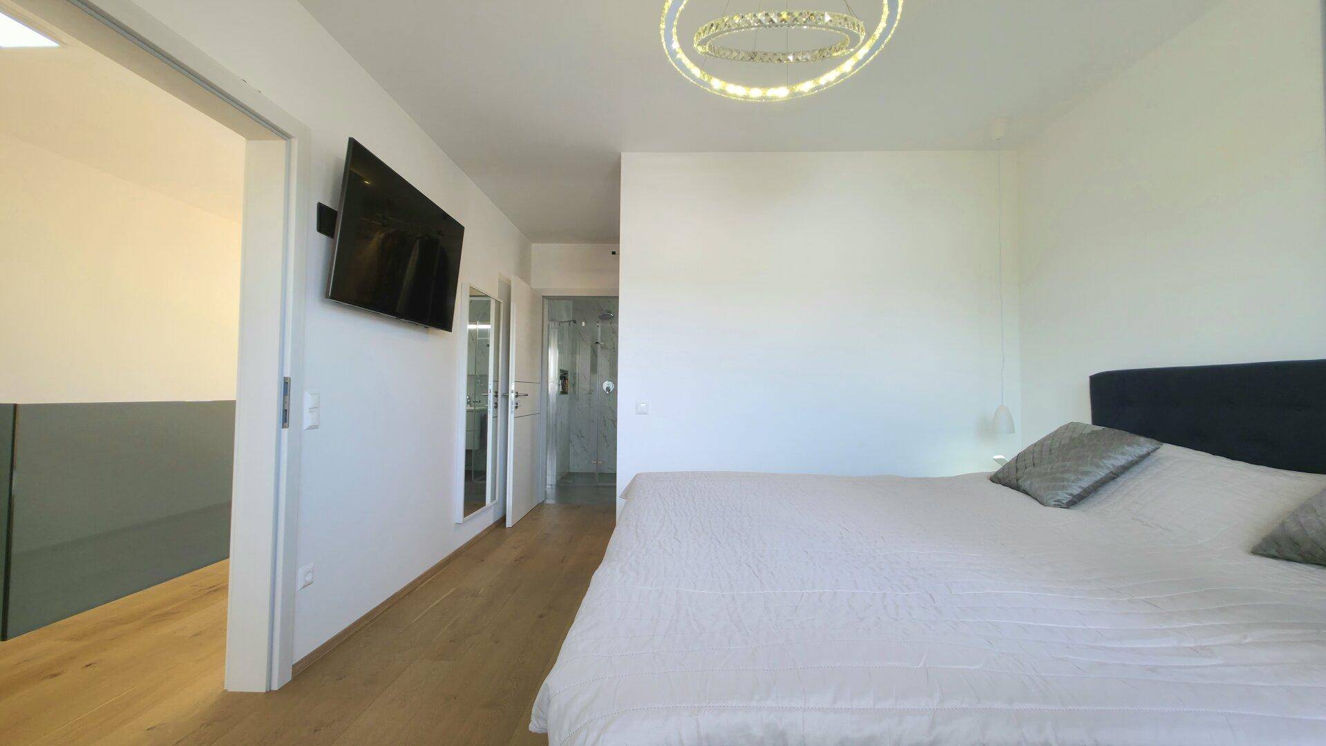 Helles Schlafzimmer mit Holzboden und Zugang zu einem angrenzenden Badezimmer, ausgestattet mit einem Fernseher.