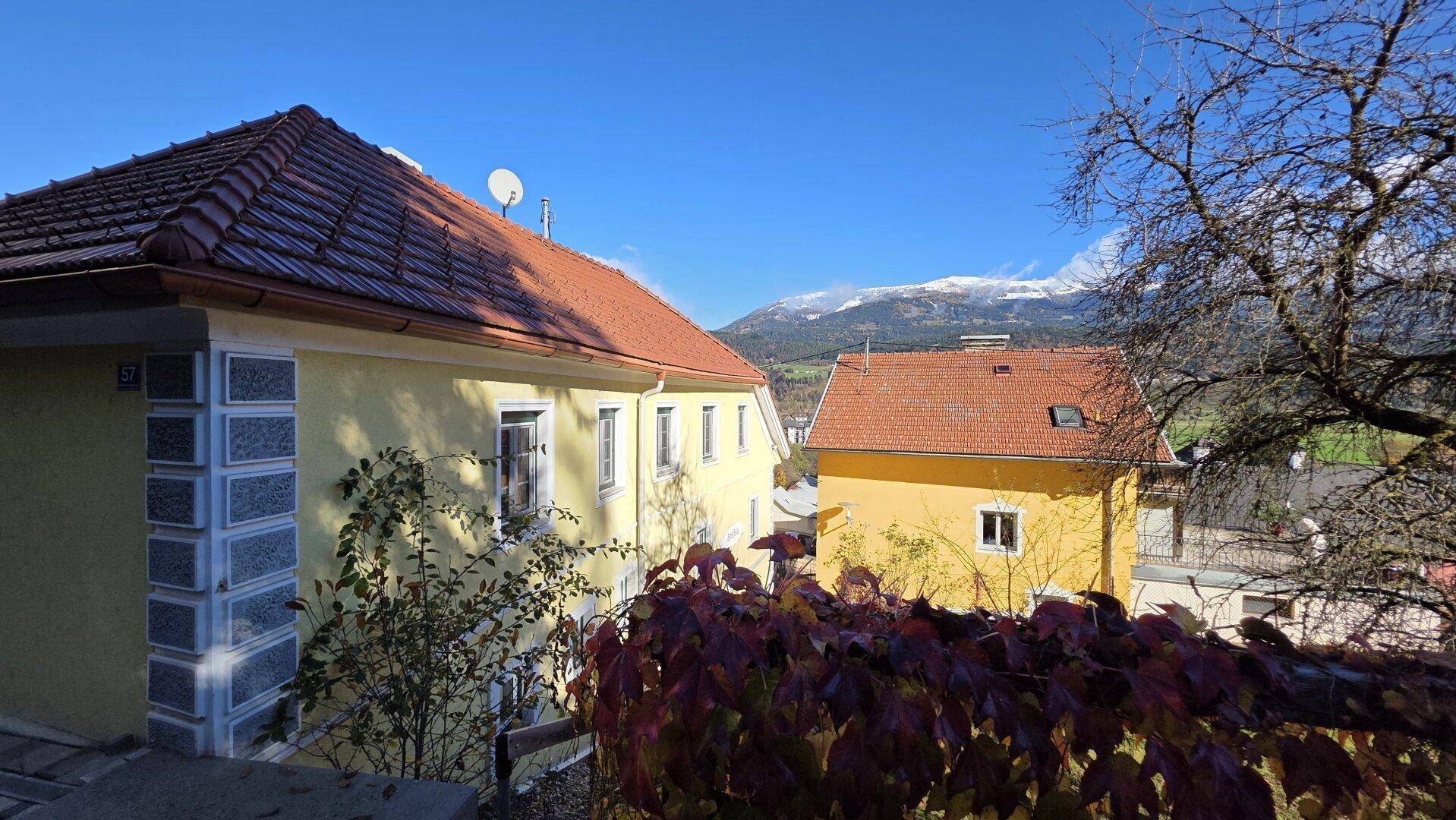 Gelbe Gebäude mit Ziegeldächern und herbstlicher Vegetation, im Hintergrund schneebedeckte Berge.