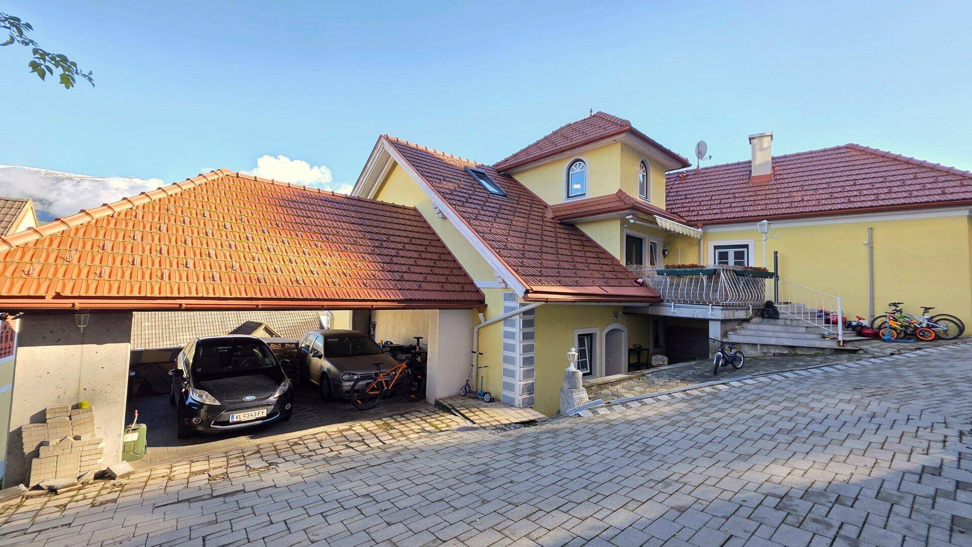 Gelbes Wohnhaus mit Ziegeldach, Carport und gepflasterter Auffahrt unter blauem Himmel.