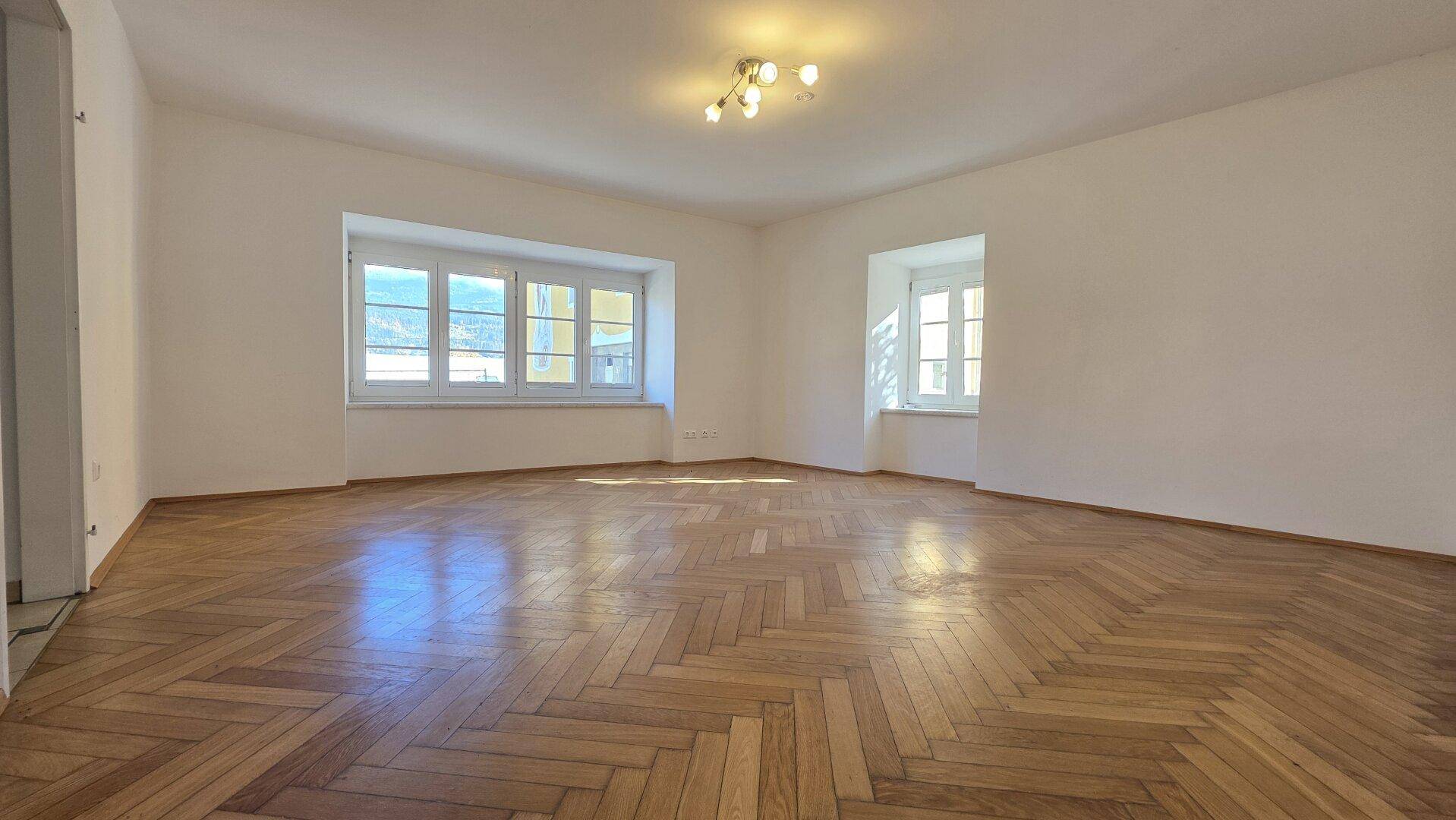 Geräumiges, helles Wohnzimmer mit Fischgrätparkett und großen Fenstern, die viel Tageslicht hereinlassen.