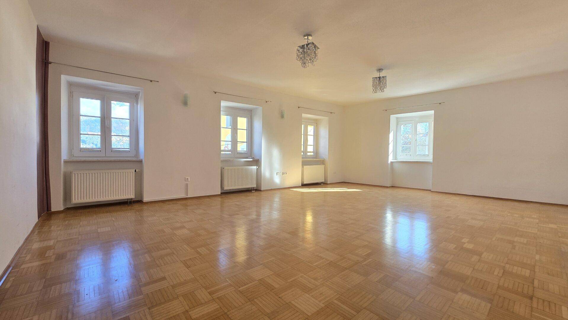 Großer, heller Raum mit Parkettboden und mehreren Fenstern, die für eine angenehme Atmosphäre sorgen.