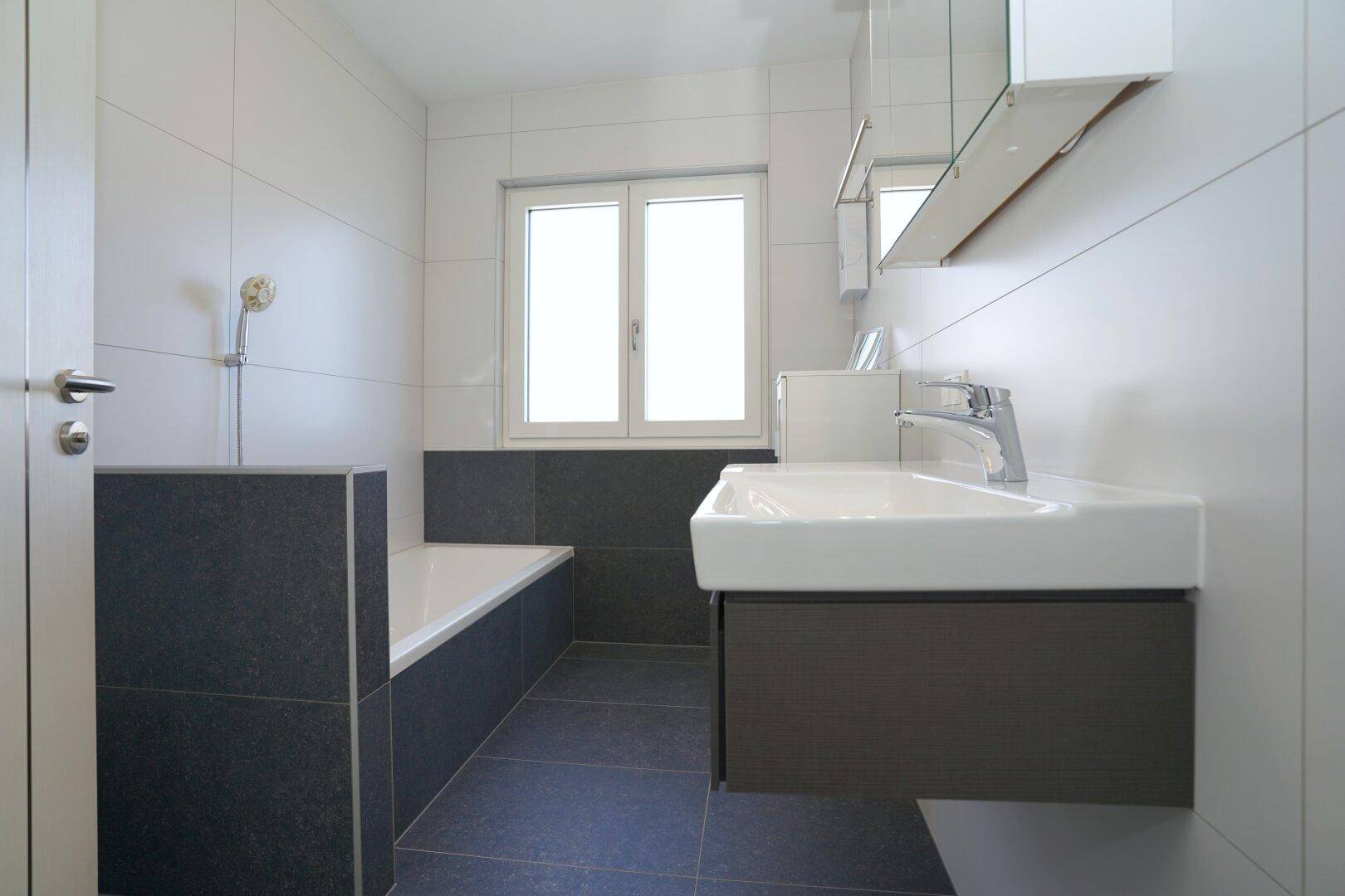 Badezimmer mit Badewanne, Waschbecken und Fenster, in schlichtem Design gehalten.