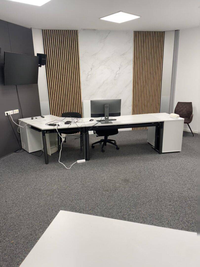Modern eingerichtetes Büro mit Schreibtischen, Stühlen und einem Wandfernseher.