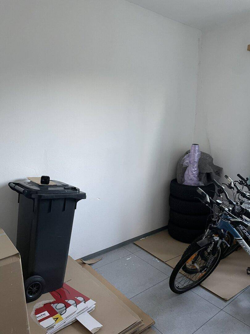 Lagerraum mit Fahrrädern, Mülltonnen und gestapelten Reifen an einer weißen Wand.