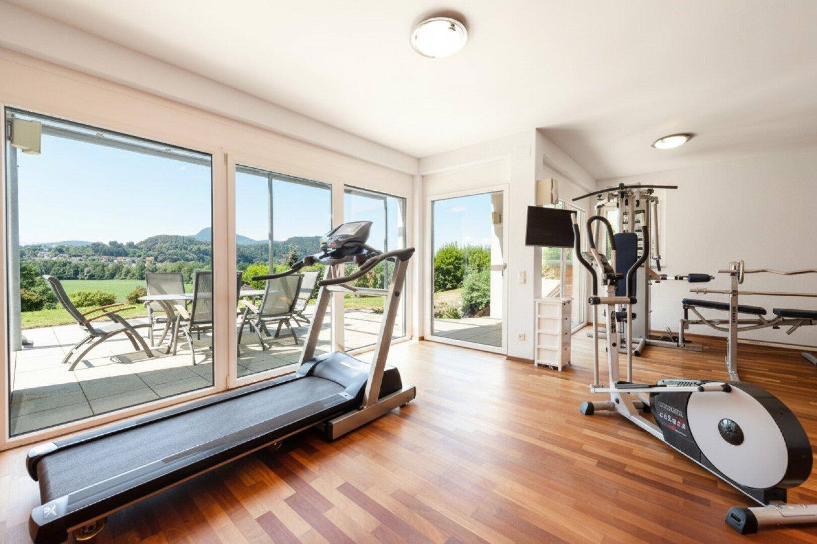 Geräumiger Fitnessraum mit Trainingsgeräten und großen Fenstern mit Terrassenzugang.