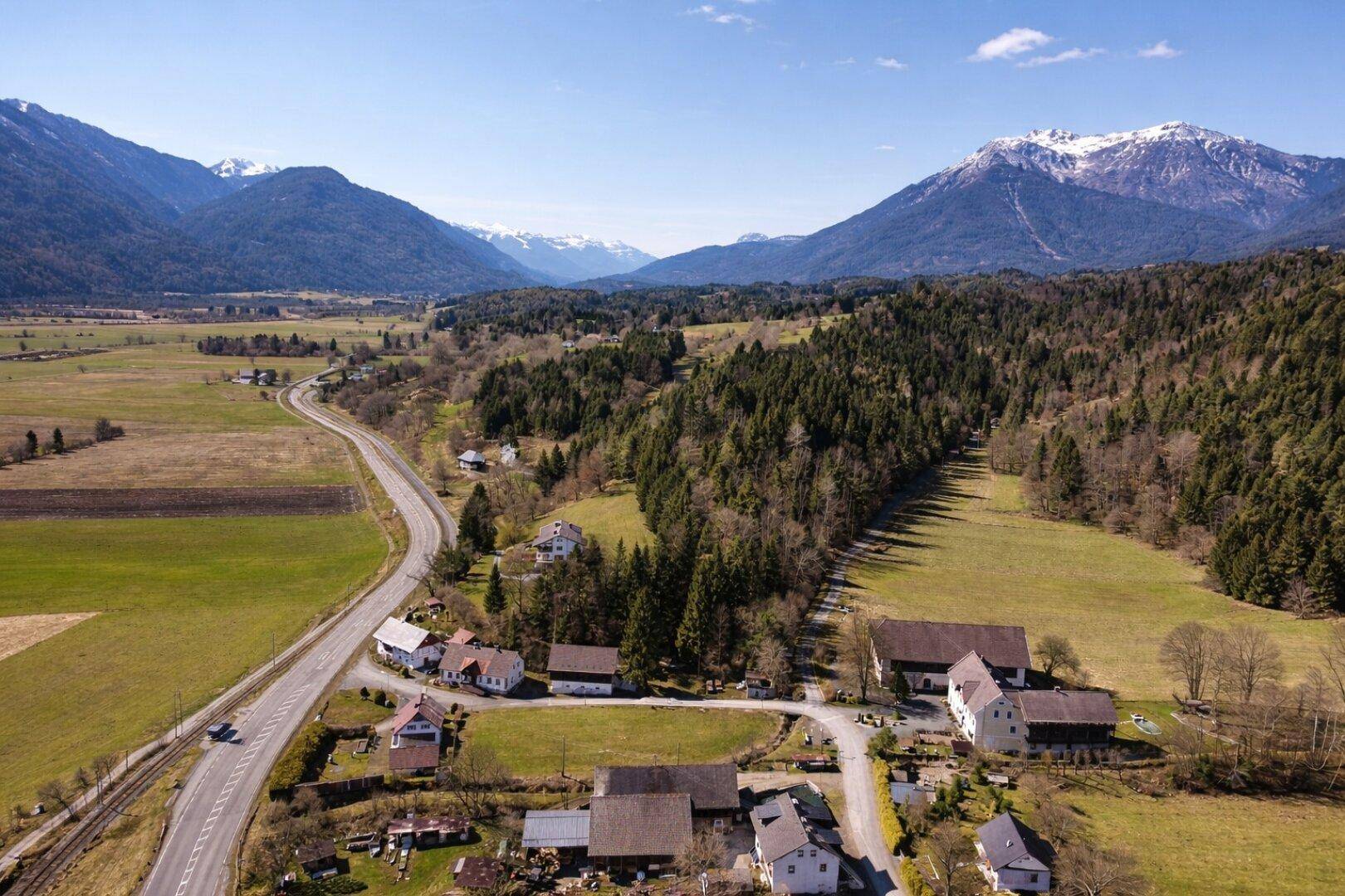 Luftaufnahme einer ländlichen Gegend mit Häusern, Feldern, Wäldern und schneebedeckten Bergen.