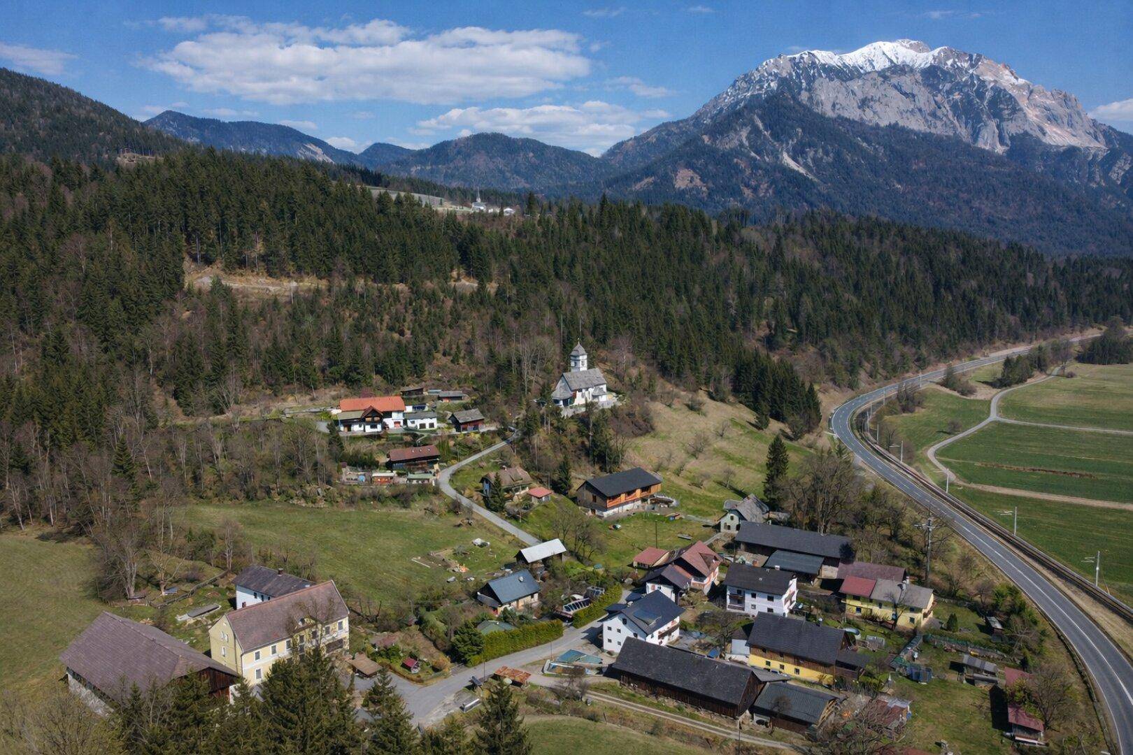 Luftaufnahme eines kleinen Dorfes in einem Tal, umgeben von Wäldern und Bergen mit einer Kirche.