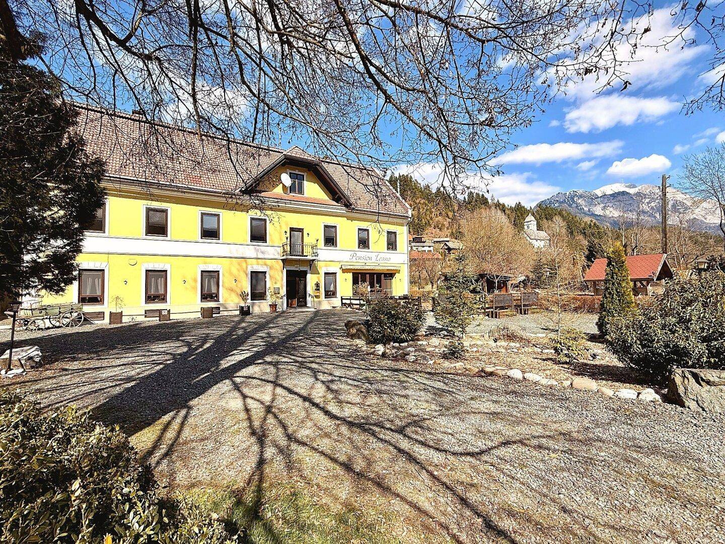 Weitläufige Ansicht der gelben Pension Leano mit großem Vorplatz und schneebedeckten Bergen im Hintergrund.