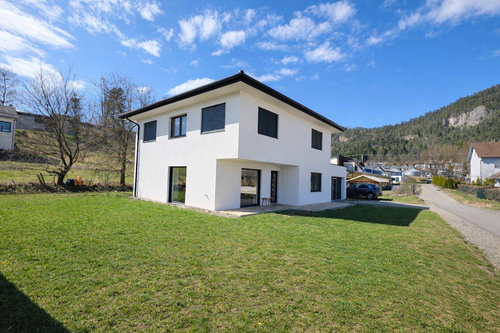 Modernes Einfamilienhaus mit weißer Fassade, schwarzen Fensterläden und großem Garten.