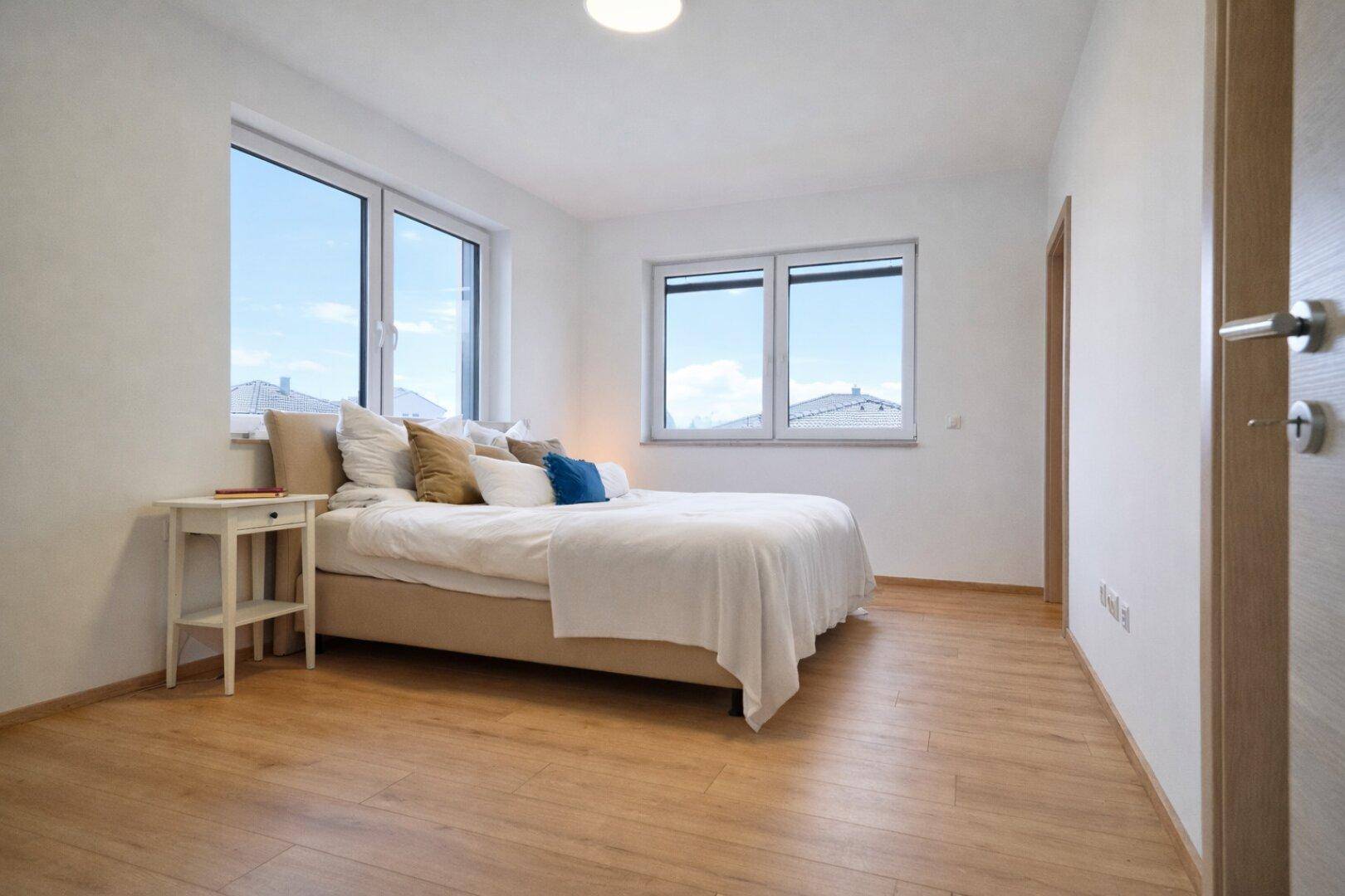 Helles Schlafzimmer mit Doppelbett, Laminatboden und Fenstern mit Blick auf die Dächer.