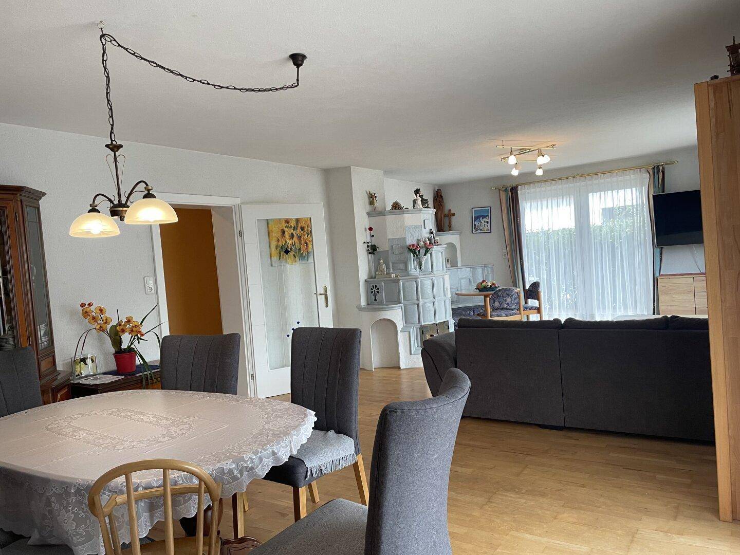 Geräumiger offener Wohn-Essbereich mit Parkettboden, Kachelofen und großen Fenstern.